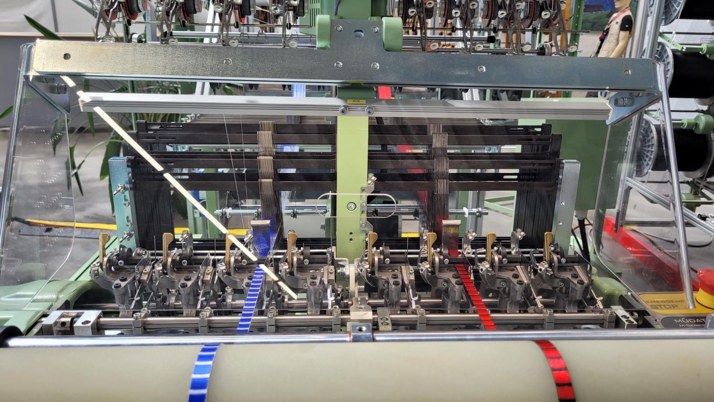 Detailansicht einer textilproduzierenden Maschine der Firma Jakob Müller aus Frick, mit sichtbaren Spulen, Fäden und mechanischen Teilen.